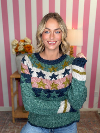 A woman wearing a green knit sweater with a colorful star pattern in navy, pink, white, and gold, posed in front of a pink and white striped wall.