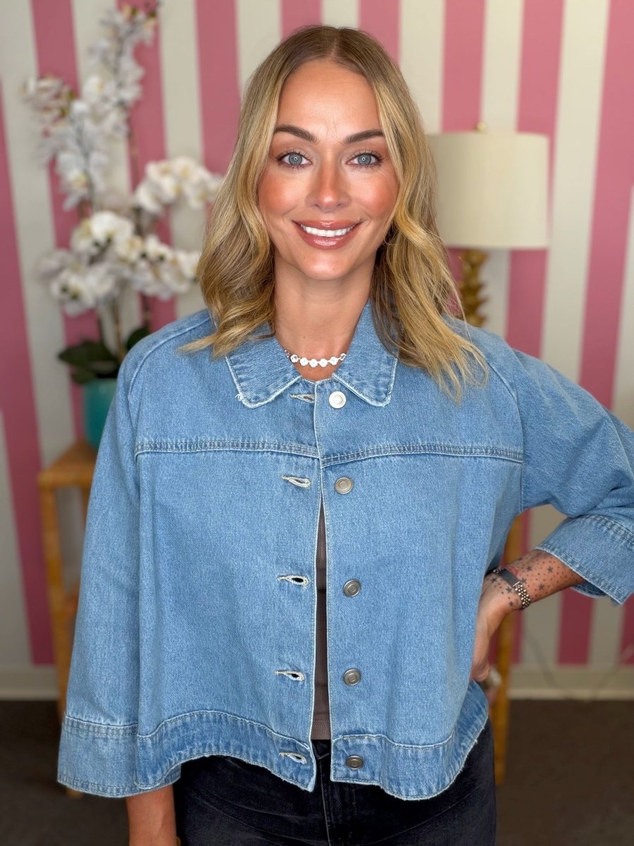 A smiling blonde woman wearing a cropped light blue denim jacket with button closures and a pointed collar, standing in front of a pink and white striped wall.