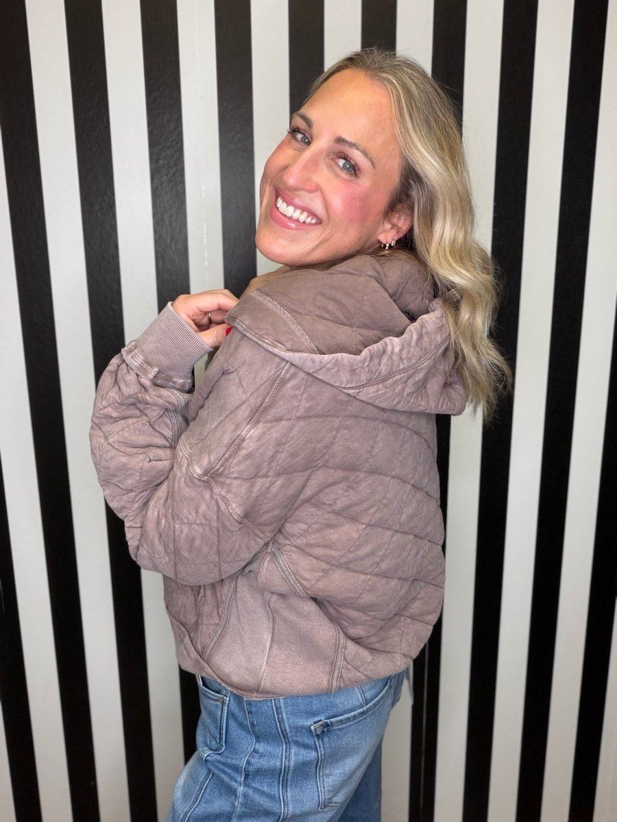 A woman with blonde hair smiling and looking back over her shoulder while wearing a crinkled, quilted taupe-brown hooded jacket and blue jeans, standing in front of a black and white vertically striped wall.