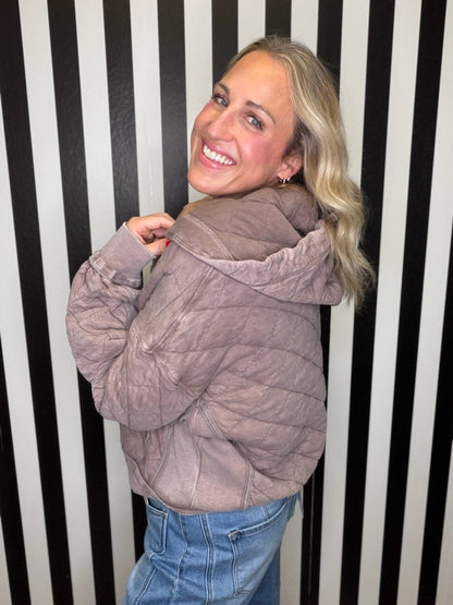 A woman with blonde hair smiling and looking back over her shoulder while wearing a crinkled, quilted taupe-brown hooded jacket and blue jeans, standing in front of a black and white vertically striped wall.