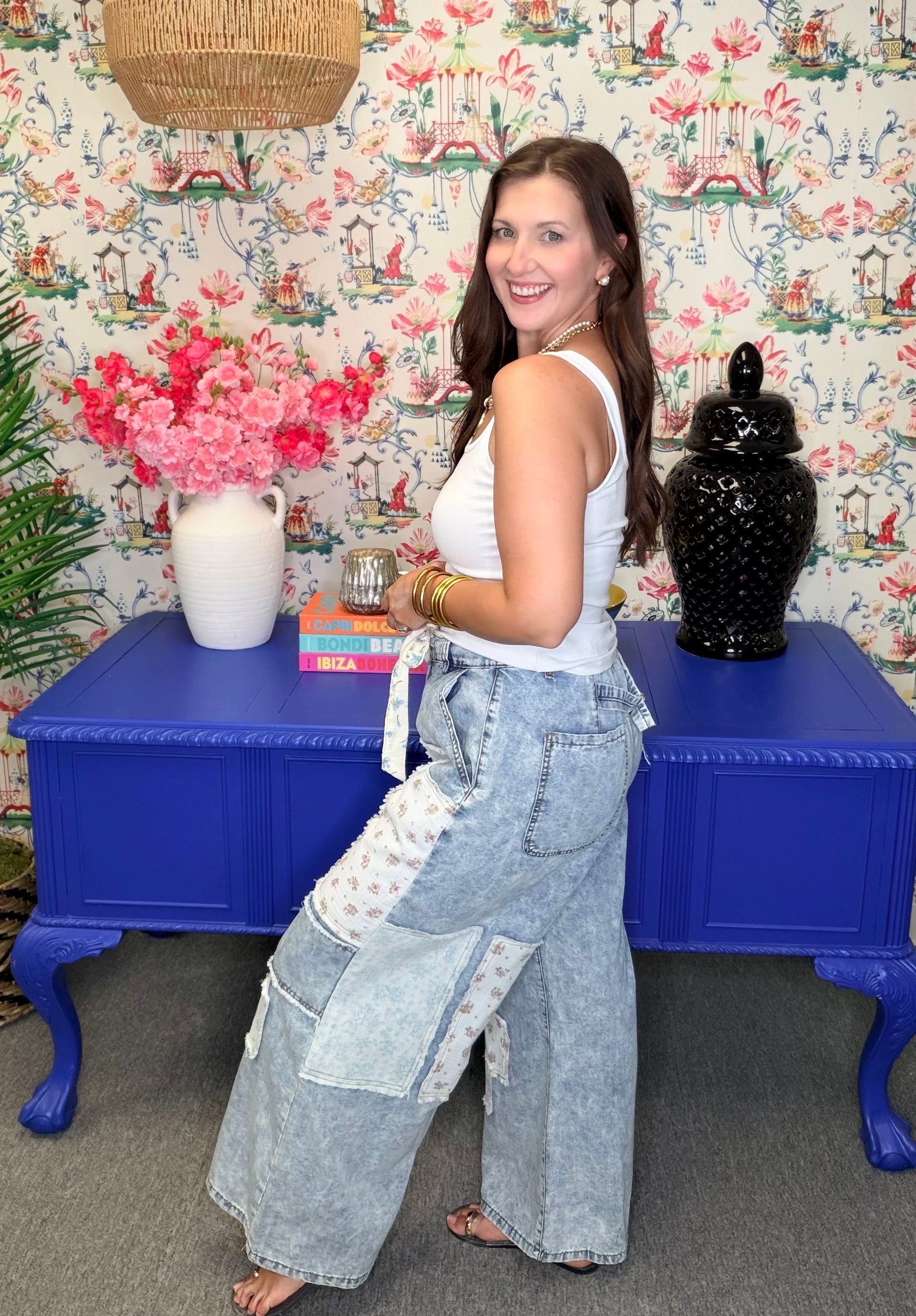 Woman in a white top and light blue pants standing in front of a colorful wall with floral patterns.