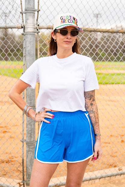 Woman wearing a colorful cap, sunglasses, white t-shirt, and blue shorts standing in front of a chain-link fence.