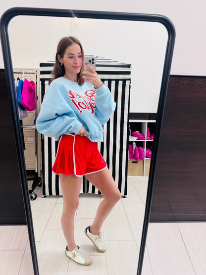 Woman taking a mirror selfie wearing a light blue sweatshirt with red text, red shorts, and white sneakers.