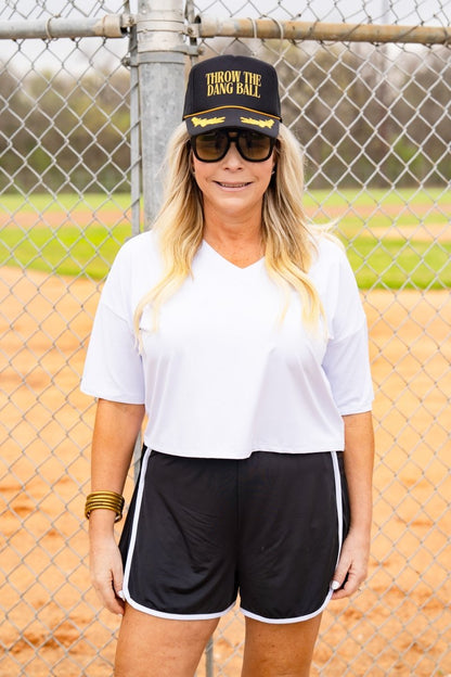 Woman wearing a white shirt, black shorts, sunglasses, and a cap with text on a baseball field.