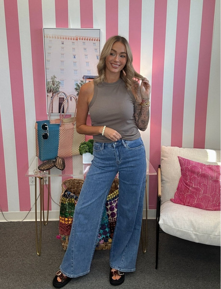 A woman stands in a room with pink and white striped walls, wearing a taupe sleeveless top and wide-leg blue jeans. She is posing next to a side table with a blue bag and a white armchair with a pink pillow.