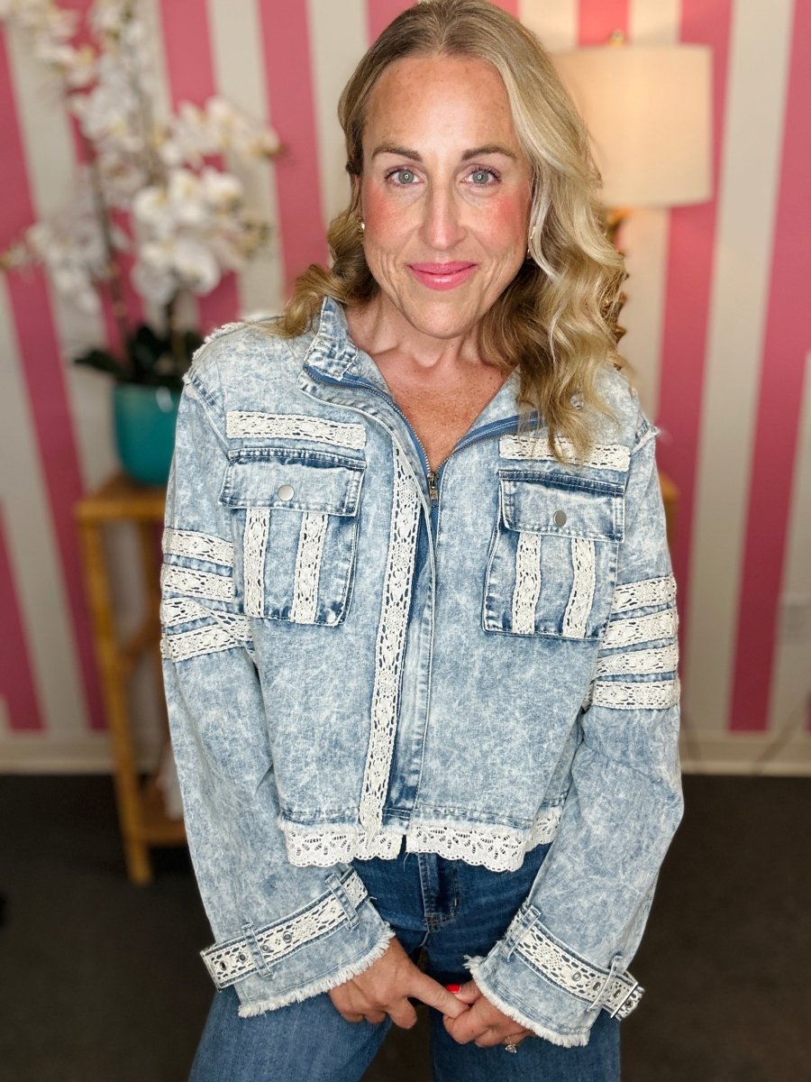 A woman wearing a light-wash blue denim jacket with white lace trim details, paired with blue jeans, standing in front of a pink and white striped wall.