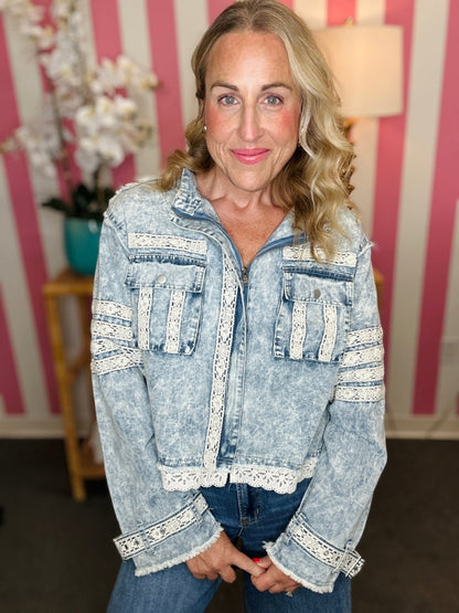 A woman wearing a light-wash blue denim jacket with white lace trim details, paired with blue jeans, standing in front of a pink and white striped wall.