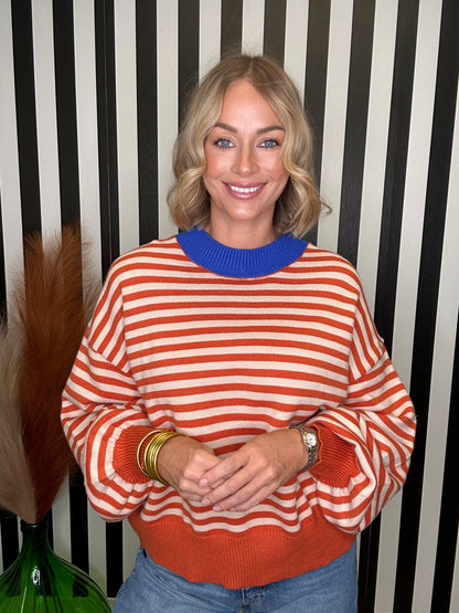 A smiling blonde woman wearing a long-sleeve knit sweater with horizontal orange and white stripes and a solid blue crew neck, standing in front of a black and white striped wall.