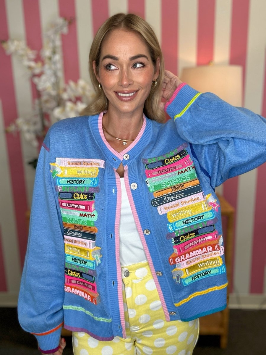 A woman wearing a bright blue cardigan jacket decorated with colorful book covers, paired with yellow polka-dot pants, posing in front of a pink and white striped wall.