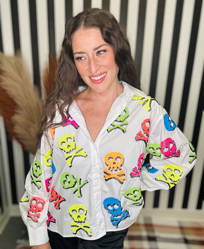 A woman wearing a white button-up shirt decorated with colorful skull and crossbones patches, posed in front of a black and white striped background.
