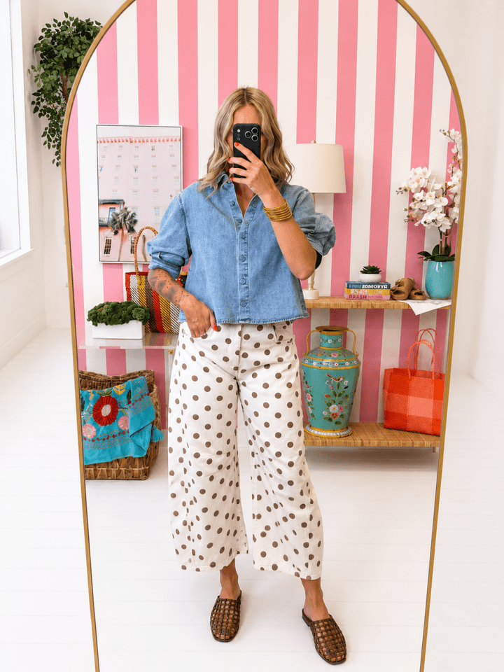 Person taking a mirror selfie wearing a denim shirt and polka dot pants in a room with striped walls.