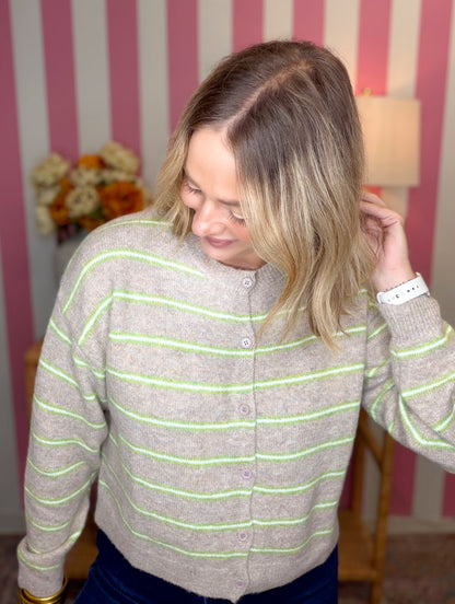 A woman wearing a beige knit cardigan sweater with thin lime green horizontal stripes, standing in front of a pink and white striped wall.