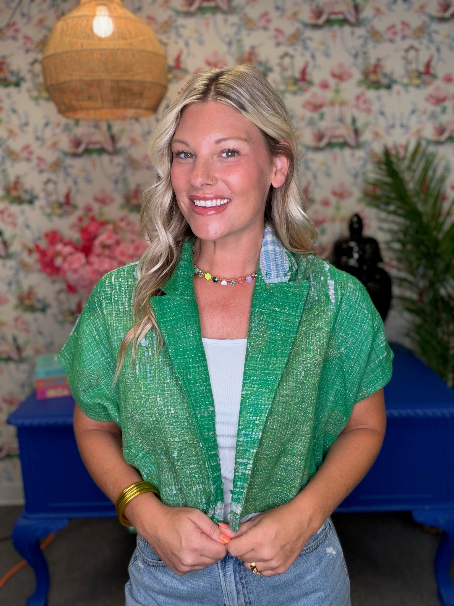 A smiling blonde woman wearing a textured green short-sleeve button-up shirt over a white top and light blue jeans, standing in front of a floral wallpaper background with a blue bench and woven pendant light.