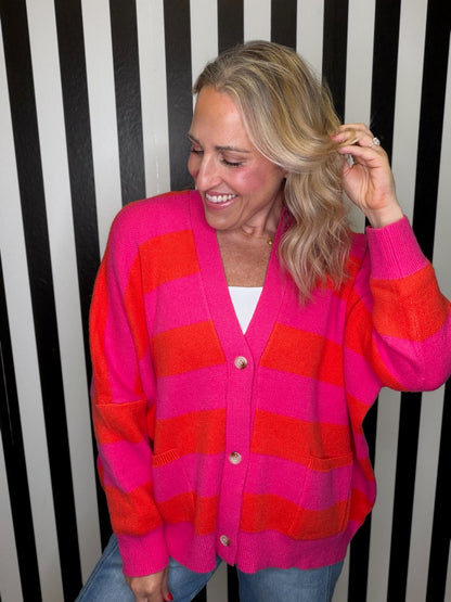 A woman wearing a bright pink and orange striped knit cardigan over a white top and blue jeans, standing in front of a black and white striped wall.
