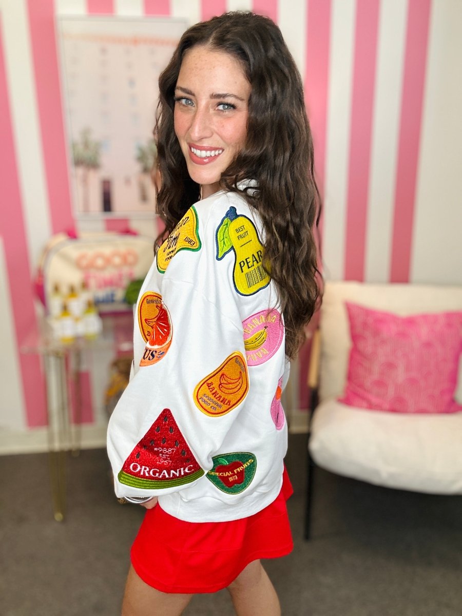 Woman wearing a white sweatshirt with colorful fruit patches in a room with pink and white striped wallpaper.
