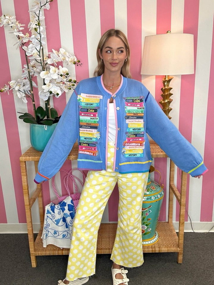 A woman stands indoors wearing a bright blue cropped jacket covered with colorful book covers, a white top, and yellow-and-white polka dot pants. She is posed in front of a pink-and-white striped wall with a lamp, flowers, and a side table.