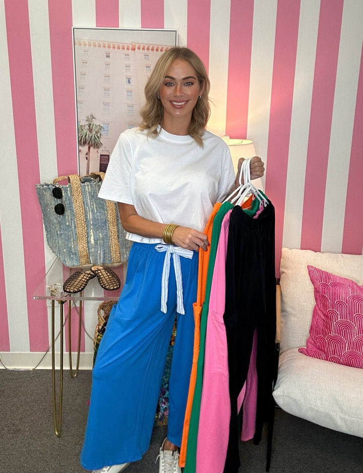 A woman stands in a pink and white striped room holding a hanger rack filled with colorful dresses, wearing a white t-shirt and bright blue wide-leg pants. A woven tote bag and a sofa with a pink pillow are visible in the background.