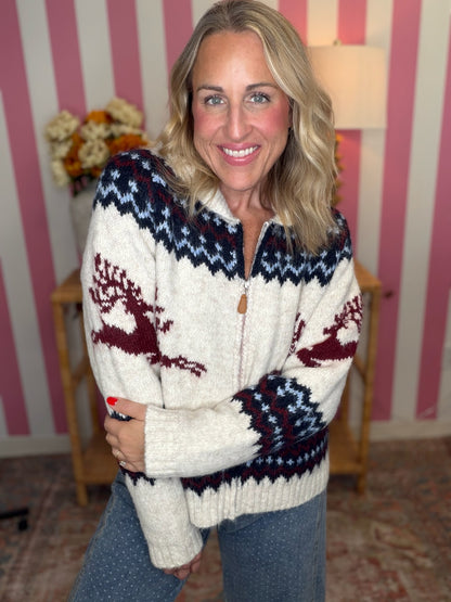 A smiling blonde woman wearing a cream knit zip-front sweater with navy and burgundy Nordic-style patterns and blue jeans, standing in front of a pink and white striped wall.