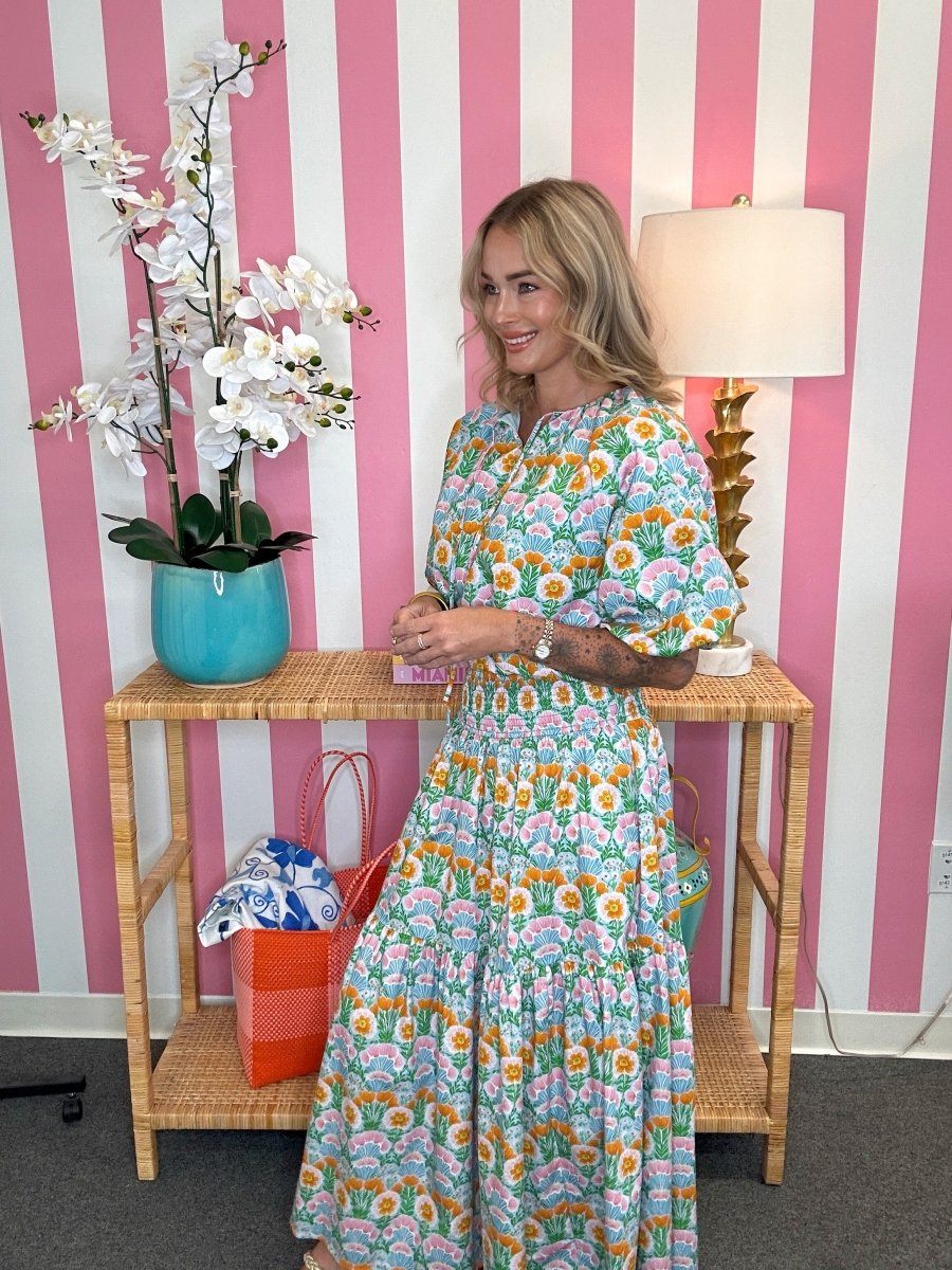 Woman in a floral dress standing in a room with striped wallpaper, a table, and decor items.