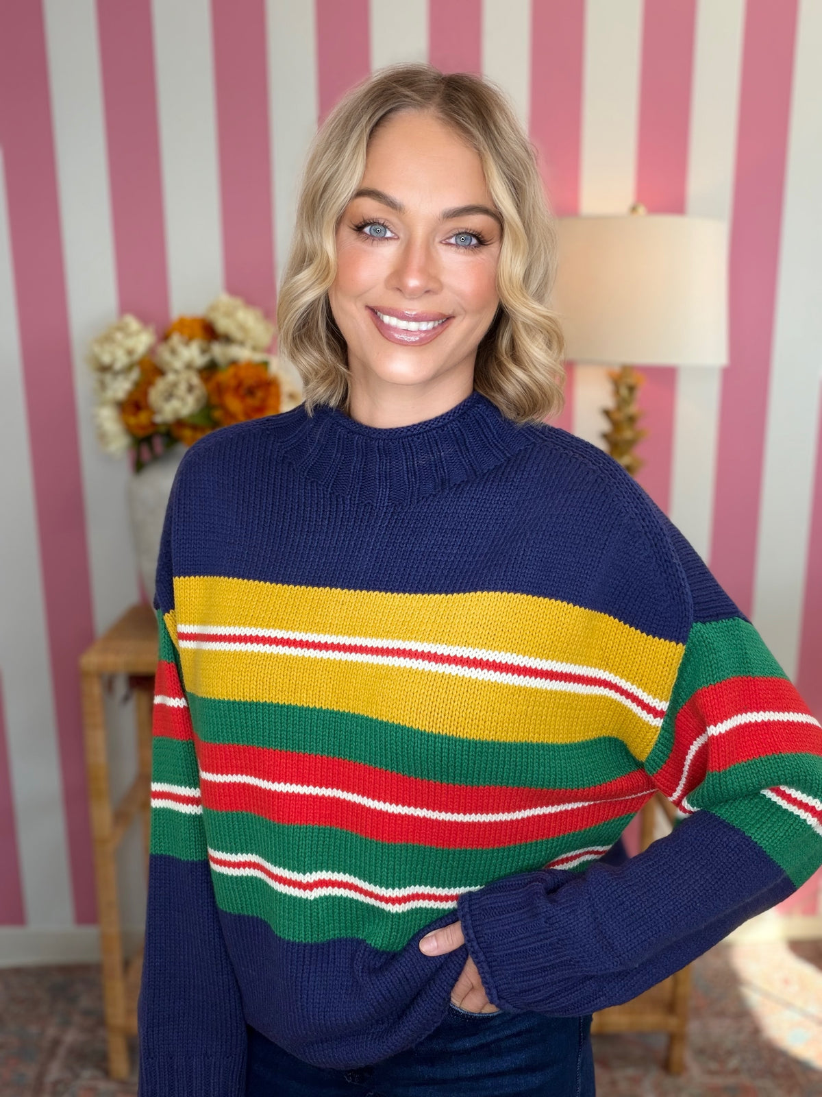 A woman wearing a navy blue knit sweater with bold horizontal stripes in yellow, green, red, and white, standing in front of a pink and white striped wall.