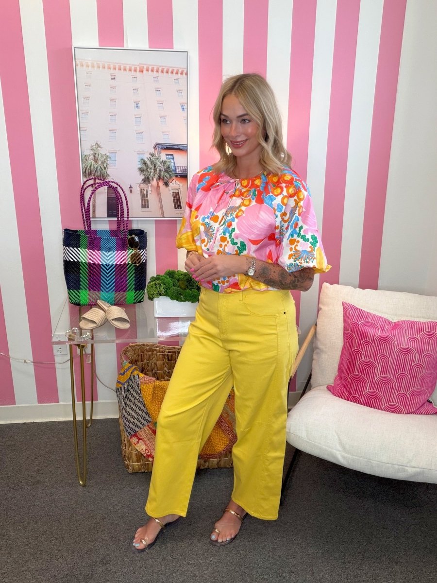 Woman in colorful top and yellow pants standing in a room with pink and white striped walls.