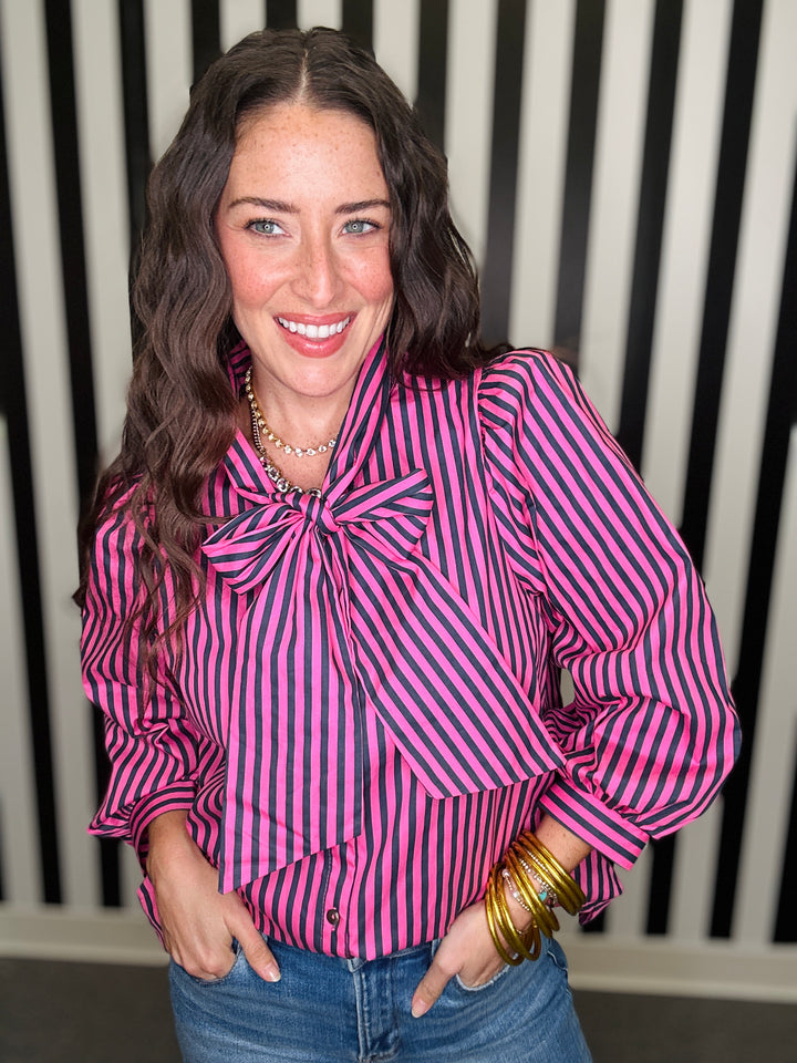 A woman smiling while wearing a pink and black vertically striped long-sleeve blouse with a large bow at the collar, paired with blue jeans and gold bracelets, standing in front of a black and white striped wall.