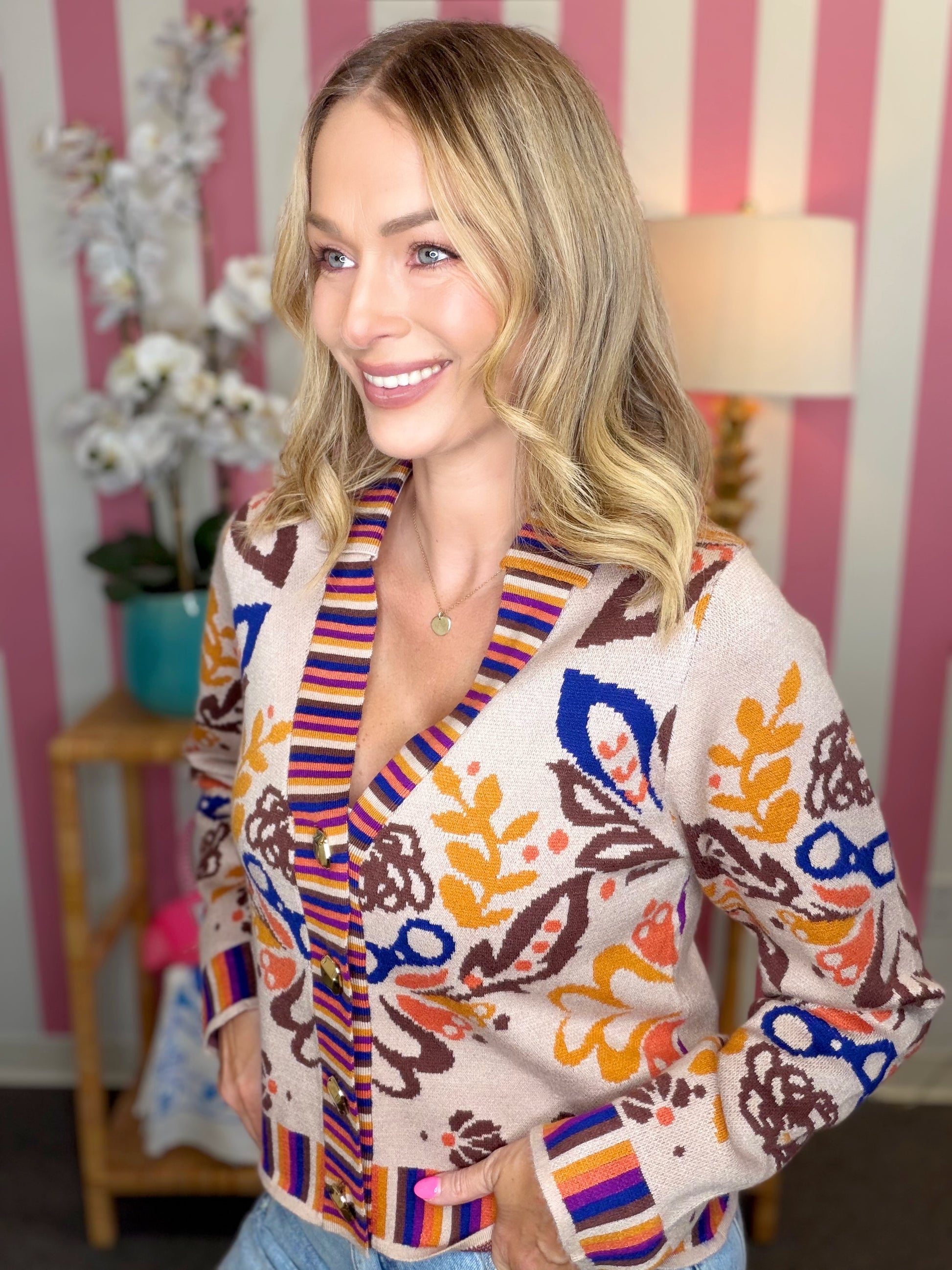 A woman wearing a cream-colored knit cardigan with colorful abstract floral patterns in blue, orange, brown, and yellow, paired with light blue jeans. She is standing in front of a pink and white striped wall with a lamp and plant in the background.