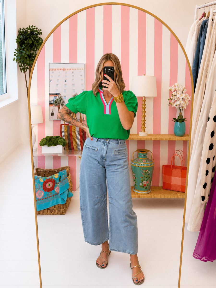 Woman taking a mirror selfie in a room with pink and white striped wallpaper, shelves with decor items, and a clothing rack.