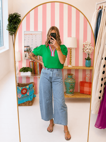Woman taking a mirror selfie in a room with pink and white striped wallpaper, shelves with decor items, and a clothing rack.