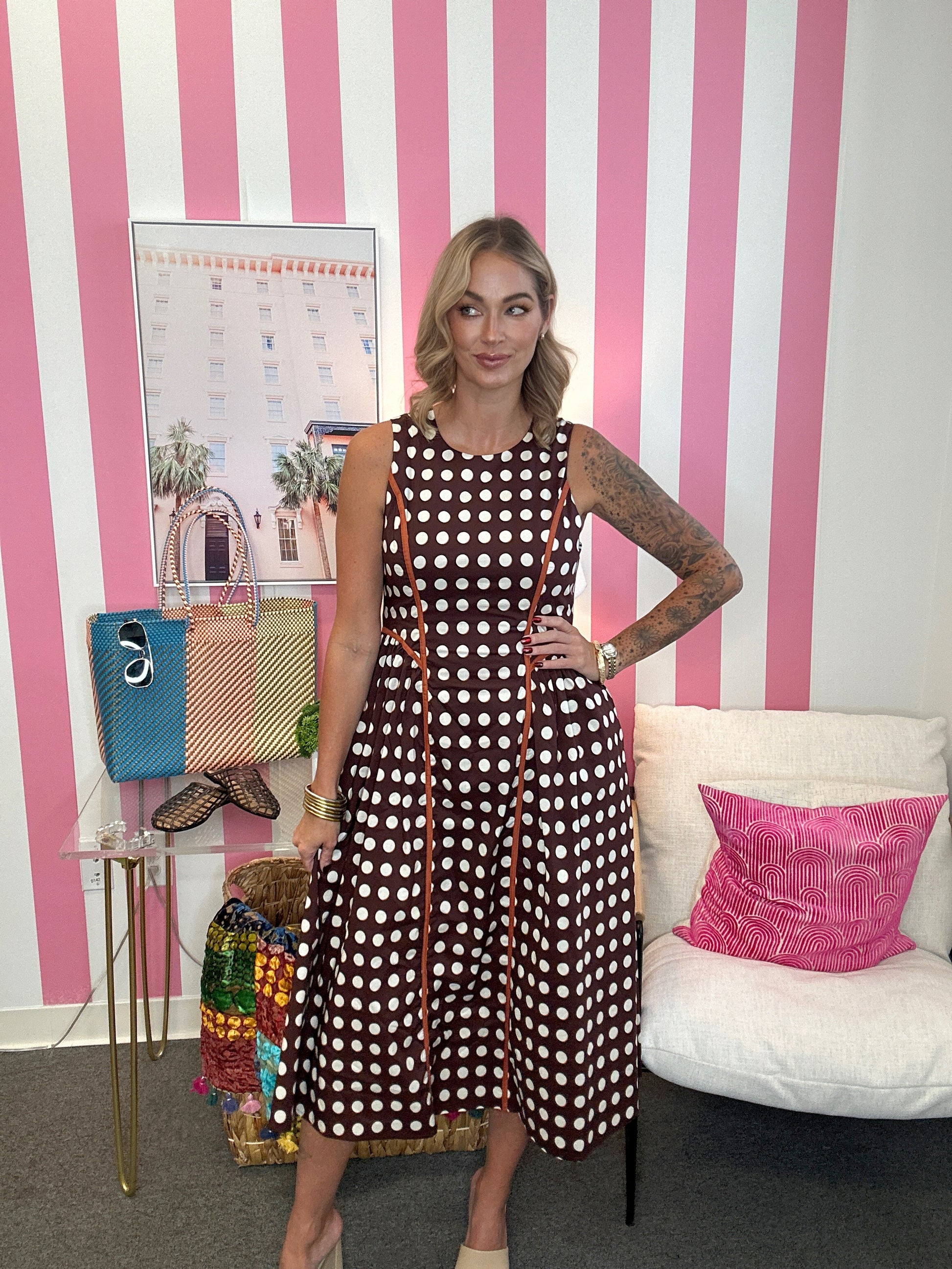 A woman models a brown sleeveless midi dress with white polka dots and orange trim, standing in front of a pink and white striped wall. She is posing with handbags on a side table and a white armchair with a pink pillow in the background.
