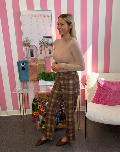 A woman posing in a beige turtleneck and brown plaid pants in a pink and white striped room with a white sofa and a glass table.