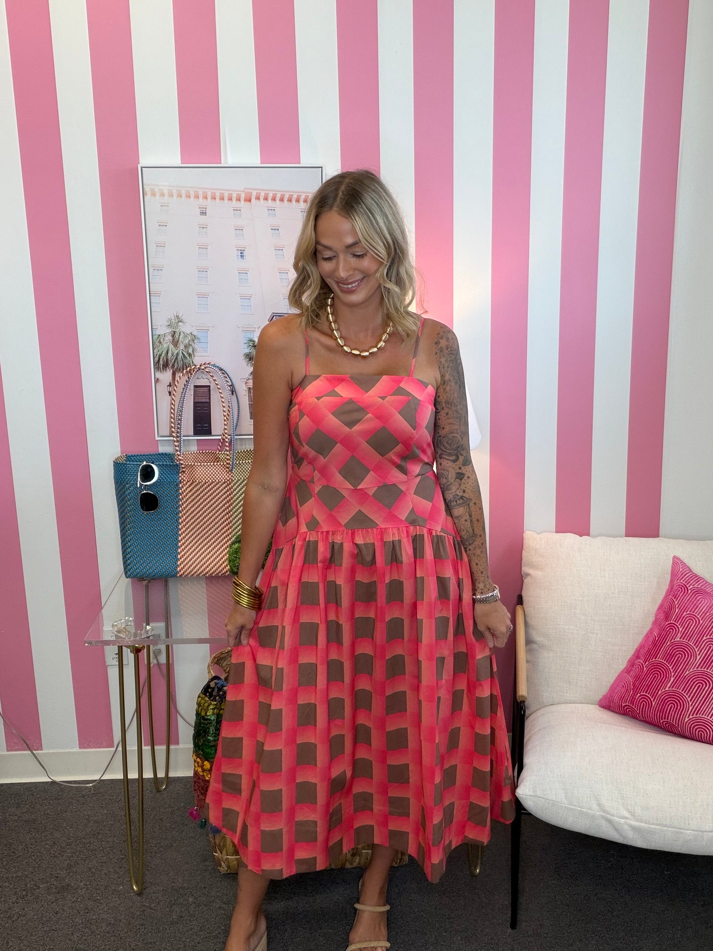 A woman wearing a pink and brown geometric patterned spaghetti-strap dress, standing in front of a pink and white striped wall.