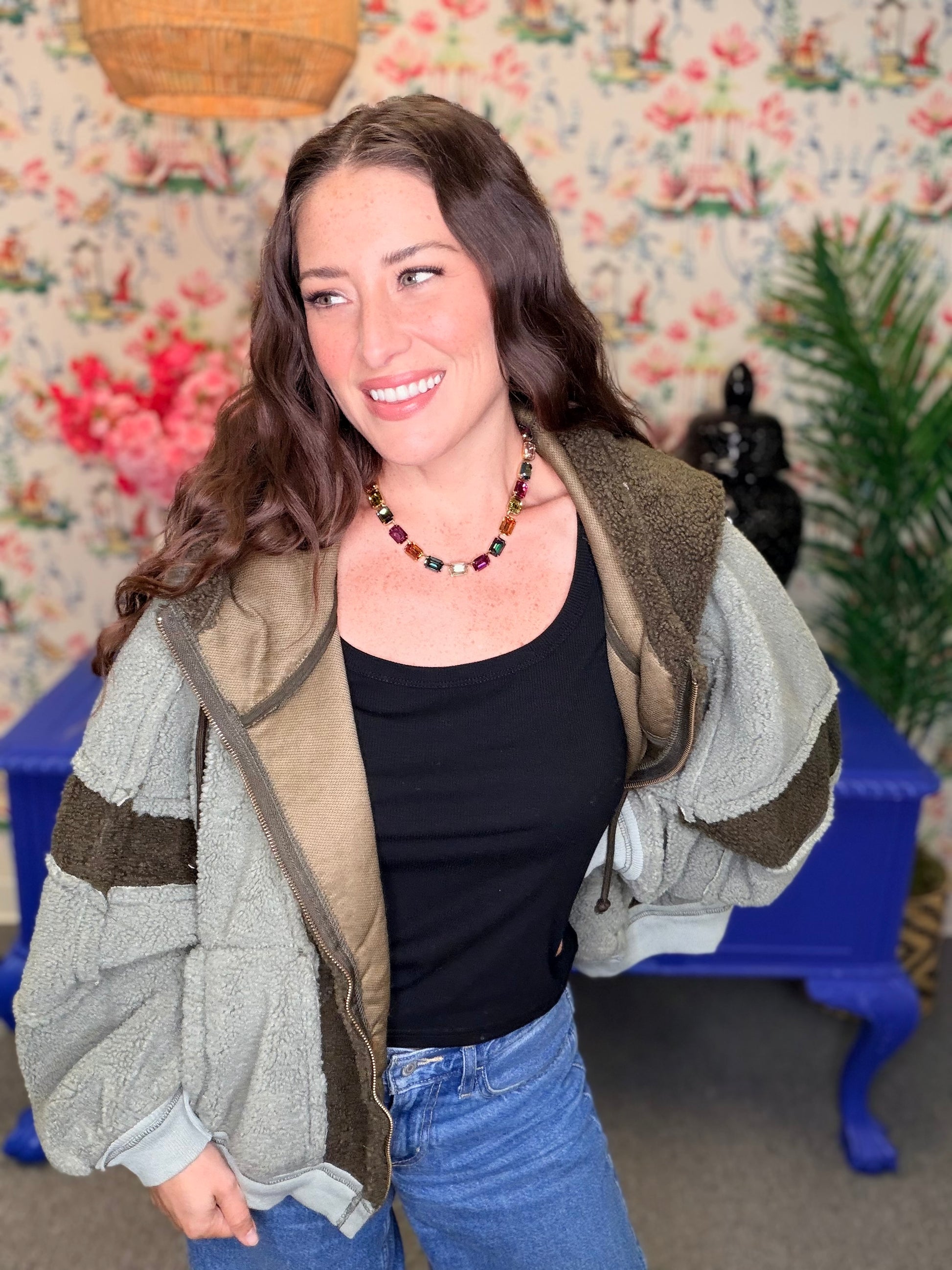 A woman wearing a light gray fleece jacket with olive green and brown striped details, a black tank top, and blue jeans, standing in front of a floral wallpaper background with a blue bench.