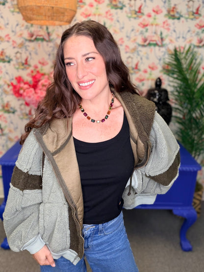 A woman wearing a light gray fleece jacket with olive green and brown striped details, a black tank top, and blue jeans, standing in front of a floral wallpaper background with a blue bench.
