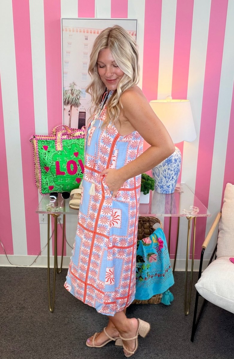 Woman in a colorful dress standing in front of a pink and white striped wall with decorative items.