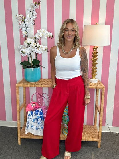 A woman stands in a pink and white striped room wearing a white tank top and bright red wide-leg pants, posing next to a wooden console table with a lamp and a potted orchid.