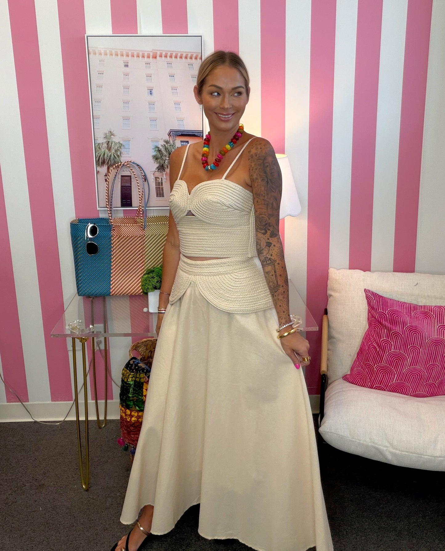 A woman stands indoors wearing a cream two-piece dress with a fitted crop top and a full skirt. The room has pink and white vertical stripes on the wall, a small table with a blue handbag, a lamp, and a sofa with a pink pillow.