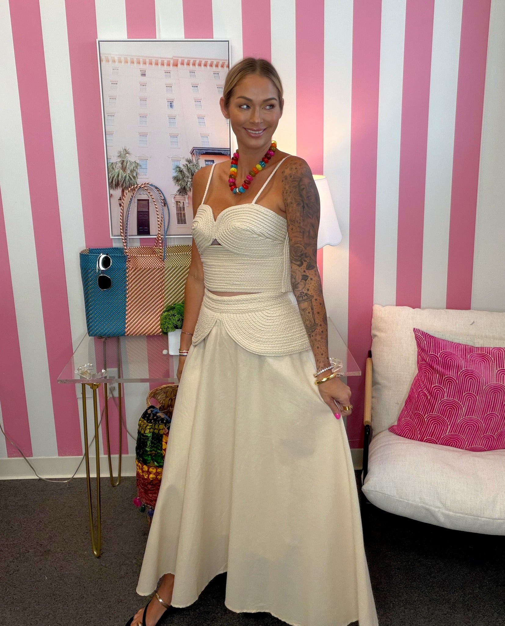 A woman stands indoors wearing a cream two-piece dress with a fitted crop top and a full skirt. The room has pink and white vertical stripes on the wall, a small table with a blue handbag, a lamp, and a sofa with a pink pillow.