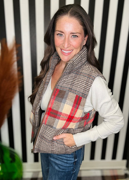 A woman wearing a brown and beige plaid zippered vest over a white long-sleeve top and blue jeans, standing in front of a black and white striped wall.