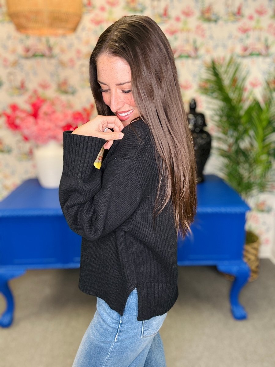 A woman with long brown hair wearing a black ribbed knit sweater and light blue jeans, standing in a room with floral wallpaper, a blue table, and plants.