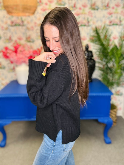 A woman with long brown hair wearing a black ribbed knit sweater and light blue jeans, standing in a room with floral wallpaper, a blue table, and plants.