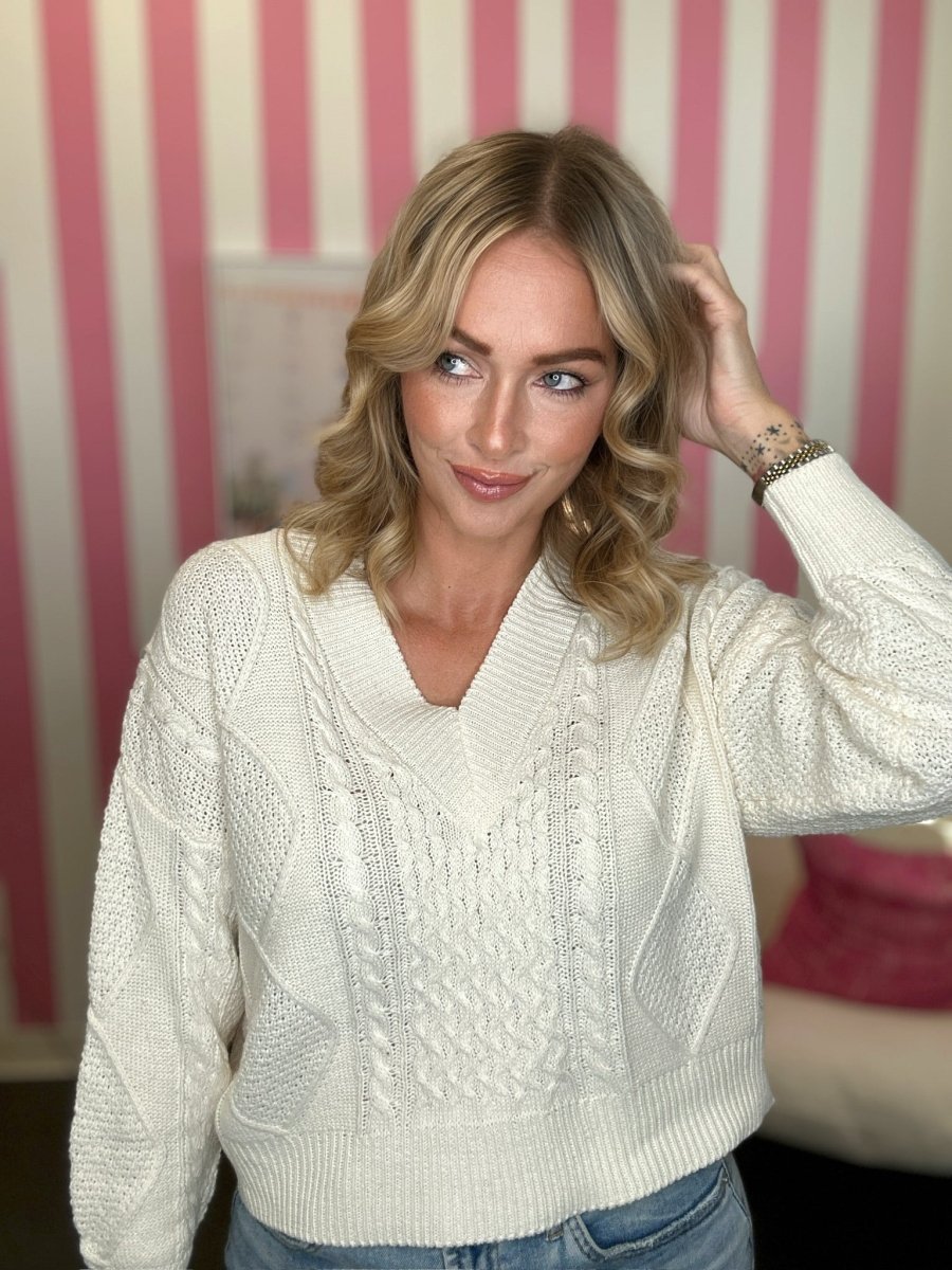 A woman wearing a white cable-knit V-neck sweater and light blue jeans, standing in front of a pink and white striped wall.