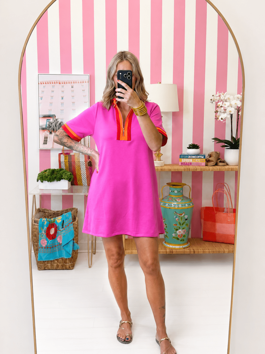 Woman taking a mirror selfie wearing a bright pink dress with orange accents in a room with striped walls.