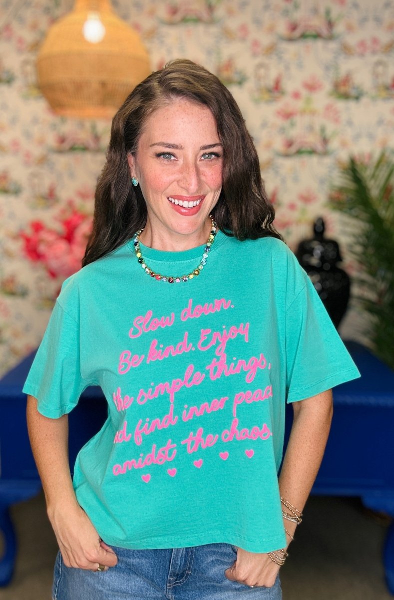 A woman smiling and wearing a turquoise graphic t-shirt with pink text that reads 'Slow down. Be kind.