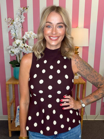 A woman wearing a sleeveless mock-neck top with a dark burgundy base and white polka dots, paired with blue jeans, standing in front of a pink and white striped wall.