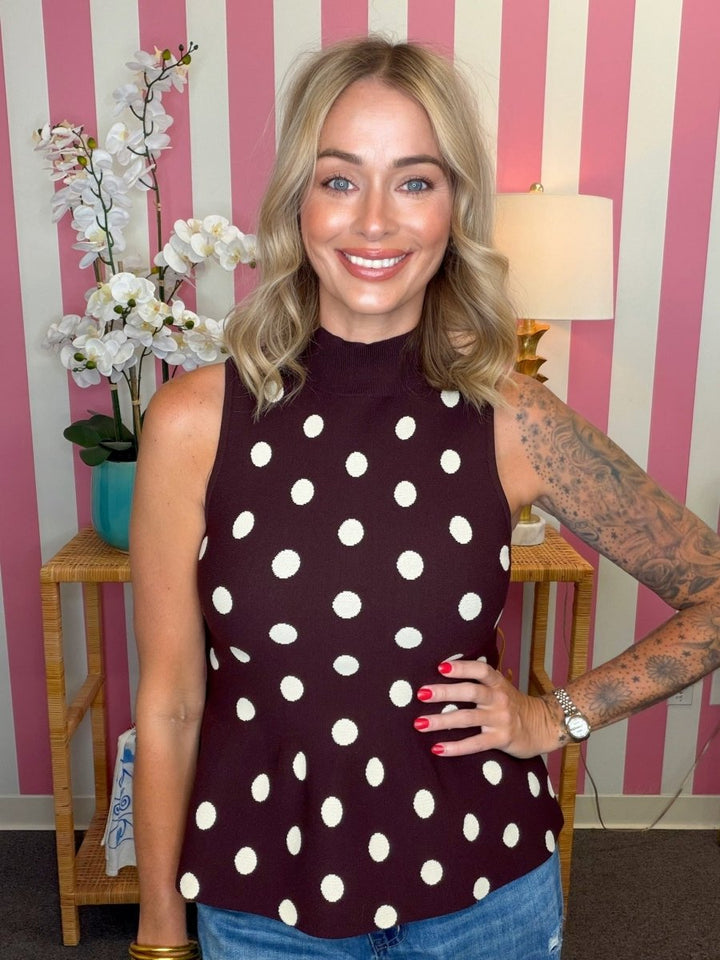 A woman wearing a sleeveless mock-neck top with a dark burgundy base and white polka dots, paired with blue jeans, standing in front of a pink and white striped wall.