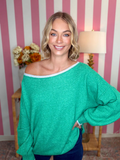 A woman wearing a bright green off-the-shoulder knit sweater with a white trim, standing in front of a pink and white striped wall.