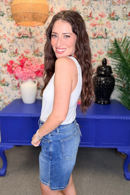 A woman with long wavy brown hair is shown from the back wearing a white sleeveless top and a blue denim mini skirt, standing in front of a blue console table and floral wallpaper.
