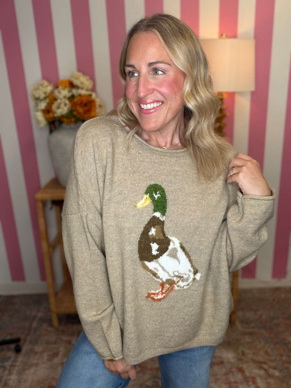 A woman wearing a beige knit sweater with a large duck motif on the front, standing in front of pink and white striped wallpaper.