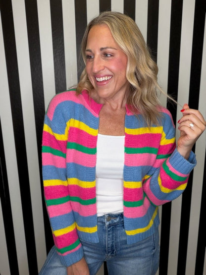 A woman smiling and wearing a colorful striped knit cardigan with pink, blue, yellow, and green horizontal stripes over a white tank top and blue jeans, standing in front of a black and white striped wall.